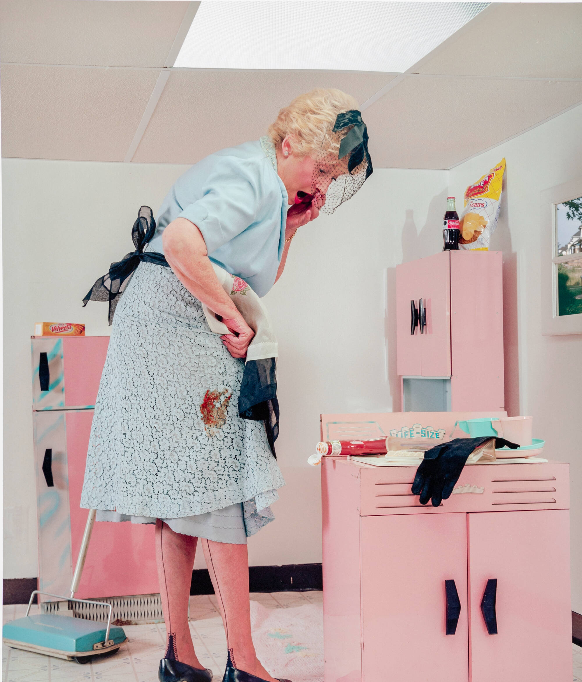 Color photograph of a woman, surprised, at a stain on her dress. She mis much larger than the pink kitchen items around her.